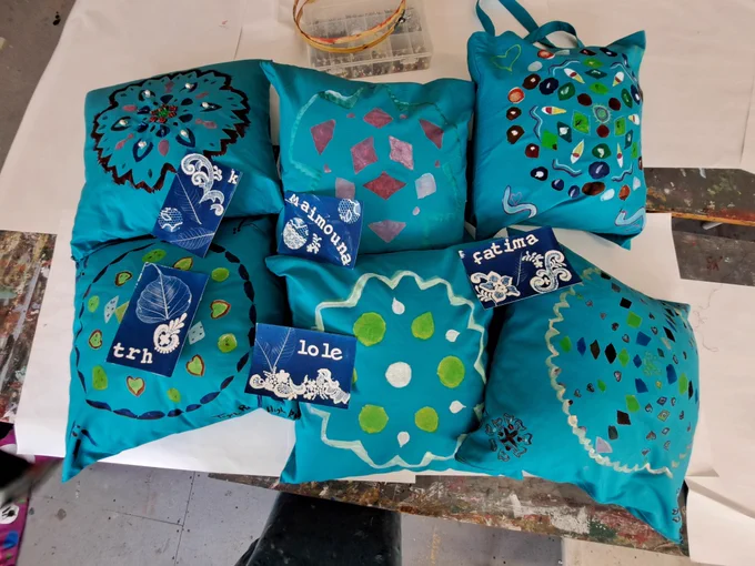 Six blue cushions with different colour print patterns and bead embroideries. They are on a white surface and on top of some of them there are small cyanotype prints with names.