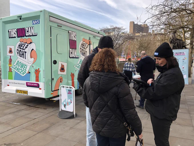A teal blue van, with cartoon images on it. Wording includes "This Van Can... help fight bowel cancer" and "Join the bowel movement - talk to us today". Images are cartoons of hands holding up placards. Location is Piccadilly gardens, Manchester. Volunteers are talking to members of the public.