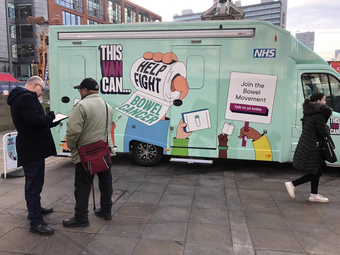 A teal blue van, with cartoon images on it. Wording includes "This Van Can... help fight bowel cancer" and "Join the bowel movement - talk to us today". Images are cartoons of hands holding up placards. Location is Piccadilly gardens, Manchester. Volunteers are talking to members of the public.