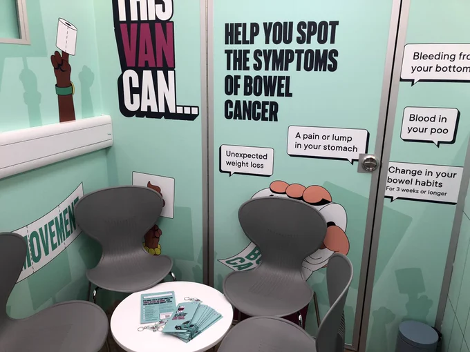 Image of inside the bowel cancer awareness van with  public health awareness messaging on the walls and leaflets and keyrings on a white table with four grey chairs.