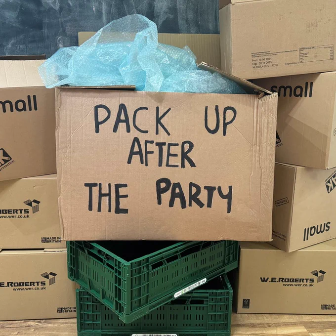 A stack of brown cardboard boxes and green crates on the floor. The box in the middle has the text ‘Pack Up After The Party’ written on it in black bold lettering, and there is blue bubble wrap coming out of the top of it.