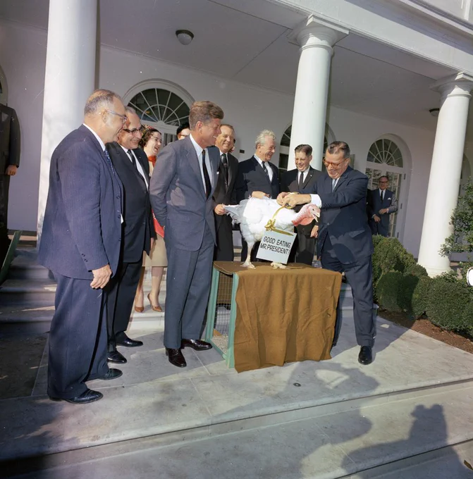 A man hangs a "Good eating, Mr. President" sign around a turkey's head while others, including President Kennedy, look on outside the White House.