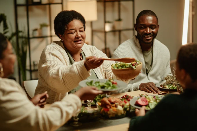 A family is seated at a table. An older woman with short dark hair wearing a sweater is holding a salad bowl and wooden spoon over the table. A man with short dark hair is seated next to her smiling. He is wearing a light green shirt under a sweater. 