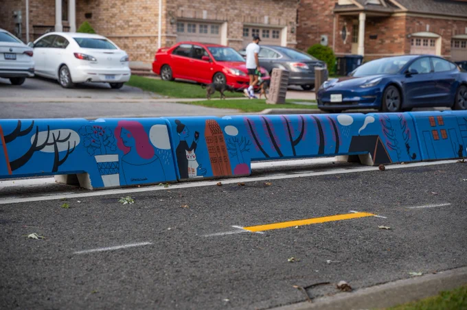 A close up photo of on eof the painted cycle track barriers installed along Ferrand Drive with artwork by artist Melika Saeeda. The artwork features images of people, pets and community against a blue background. Behind hte cycle track a person walking a dog can be seen walking along the sidewalk
