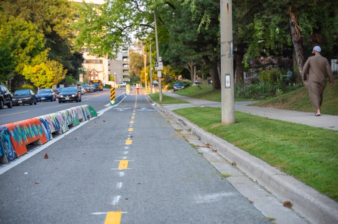  A photograph of vibrant cycle track barriers, installed along Ferrand Drive. A pedestrian can be seen walking on the sidewalk on the right hand side of the image.