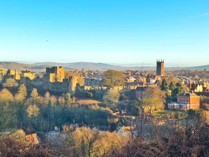 A photograph of the town of Ludlow in bright winter sunshine under a shockingly blue sky, with the castle to the left and the church to the right and the houses of the town nestled between. The Shropshire Hills recede into the distance behind. 