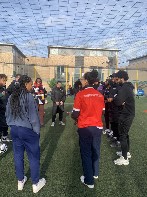A group of around 12 people are stood in a circle on an outdoors artificial grass football pitch in front of a light brown building and blue sky. The closest person is stood with their back to the camera and is wearing a red shirt with "FOOTBALL ON THE BRAIN" in white capital letters across the back.