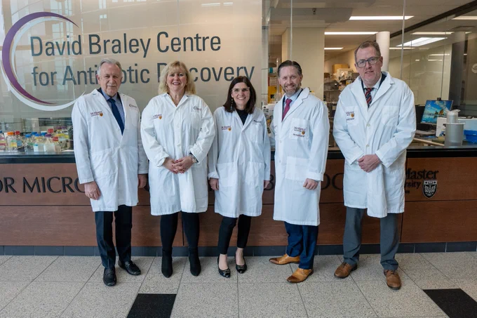 Five people, all wearing white lab coats that have a McMaster University logo on them, smile directly at the camera. Behind them is a sign that reads, ‘David Braley Centre for Antibiotic Discovery.’
