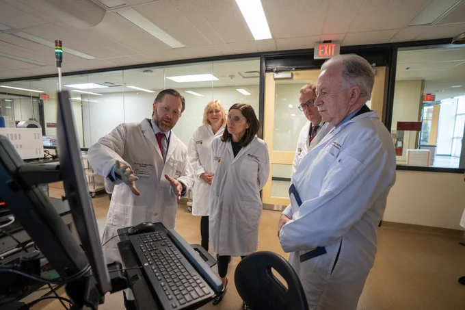 A researcher explains something while pointing to a computer screen. Four other people stand around him listening. They are all wearing white lab coats with the McMaster University logo on them. 