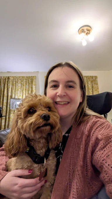 Me smiling in my wheelchair with a pale brown dog (cavapoo) on my lap 