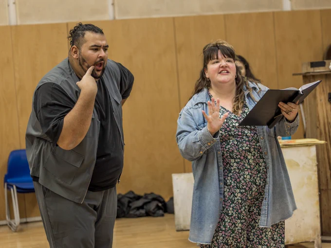 Marc Zayat is Ensemble and Jamie-Rose Monk is Ensemble in Hansel and Gretel (2024/5). 

Two actors rehearse in a rehearsal studio. On actor reads from a binder they hold in front of themself. The other actor looks over their shoulder. 