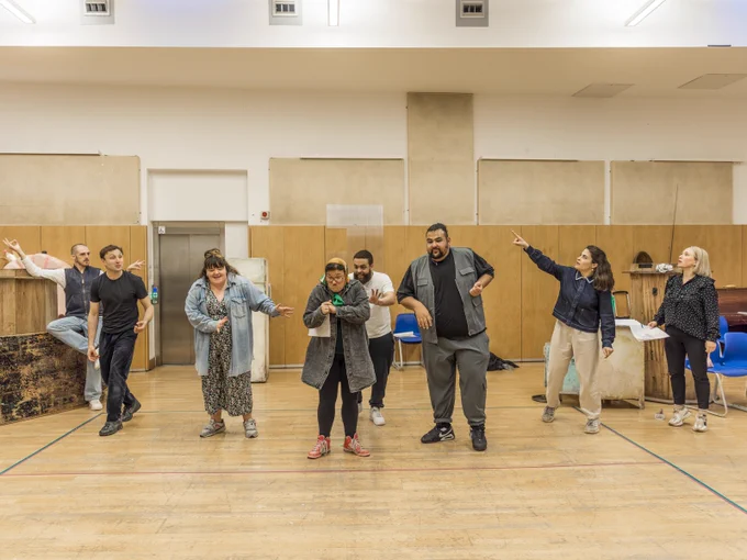 A group of actors rehearse in a rehearsal studio at the Globe. The all stand in a line, practicing choreography. 