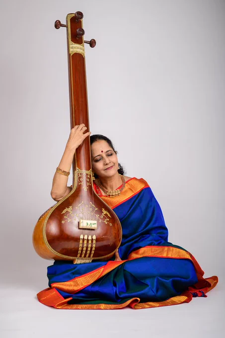 Portrait of a singer. It's a studio shot on a white backdrop. She is sitting on the floor, holding her tanpura vertically on her lap. She is leaning her head on the neck of the tanpura and her eyes are closed. She is wearing a deep blue sari with a red and gold border.