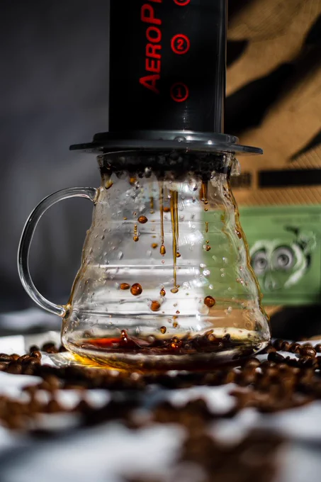 A photograph of an Aeropress, in the process of pressing coffee into a glass carafe. There are droplets of coffee falling into the carafe and a stream falling out of the Aeropress. Behind the carafe is a bag of Black Baza coffee called Loris. This was shot for Black Baza Coffee.