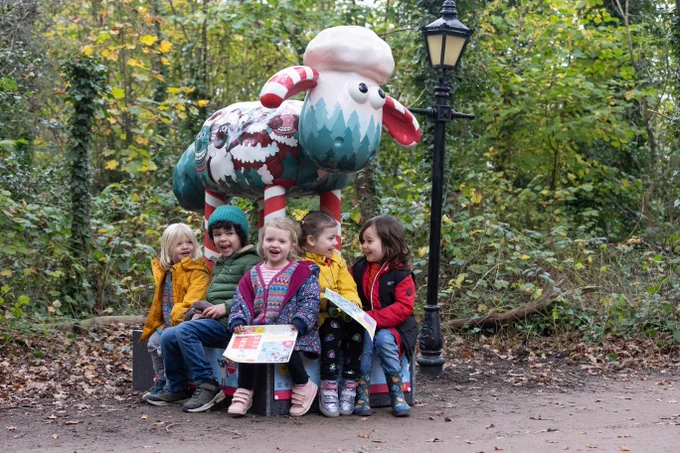 Five children are sitting and smiling in front of a large, colourful sculpture of Shaun the Sheep. A lamp post stands to the side in a woodland setting with autumn leaves.