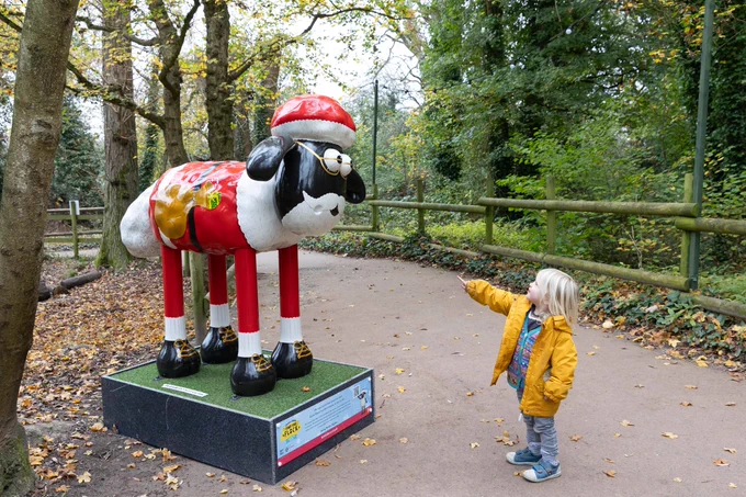 A child interacts with a large sculpture of Shaun the Sheep dressed as Santa Claus, located outdoors in a park-like setting.