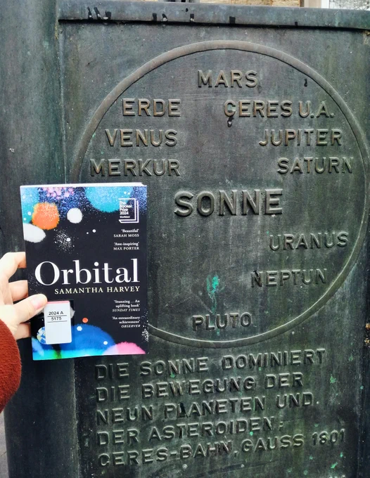 Holding the book "Orbital" next to a plate listing the sun and the planets of the solar system
