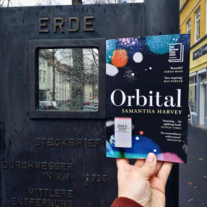 Holding the book "Orbital" next to the artistic rendition of "Earth" as a tiny metal ball between two sheets of glass
