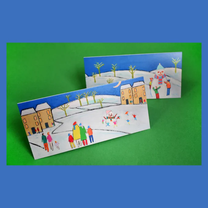 A blue background for a photo of two cards. The cards are placed on a green backdrop and have different designs of children playing in the snow and decorating snowmen. The artwork is by David Bradley.
