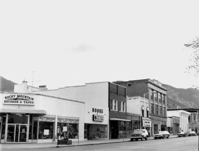 Black and white photo of the 1300 block of Pearl Street. Rocky Mountain Records & Tapes is seen on the lefthand side. Window signs advertise types of music including Bluegrass, Folk, Rock, and Blues. A few cars line the street