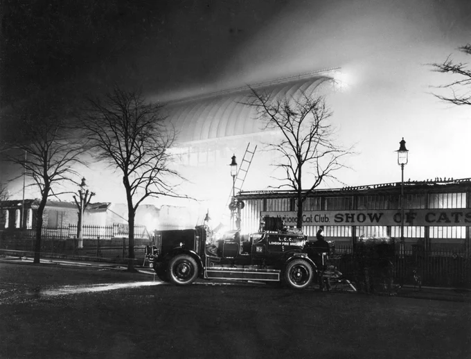 A black and white photo showing a London Fire Brigade fire engine parked in front of Crystal Palace. The frame of the glass building is enveloped in smoke and flames. 