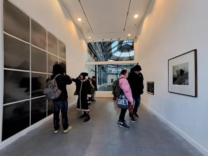 A group of people stand in a narrow gallery space. There is a big window at the end of the room and dark, black and white artworks hung in the white walls. Everyone is dressed for the cold weather.