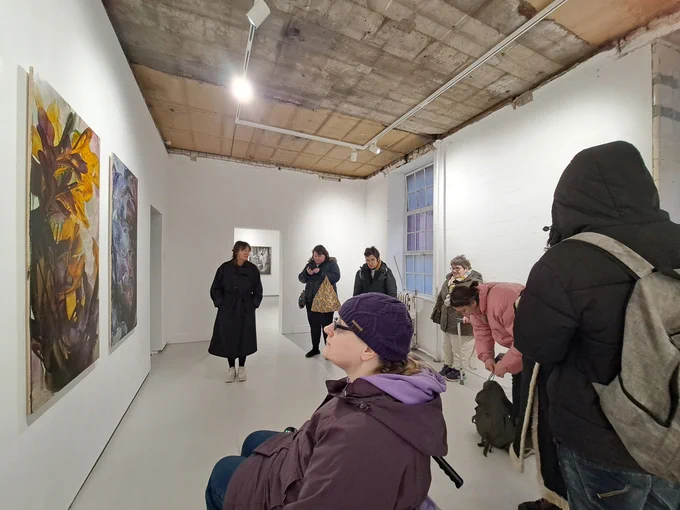A group of people gather in a spacious gallery with white walls. There are two big artworks of flowers on the wall.