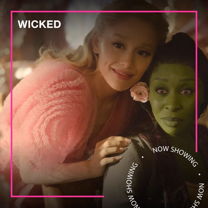 A woman in a pink frilly dress and a woman with green skin stand together hugging. Text - Wicked, Now Showing

