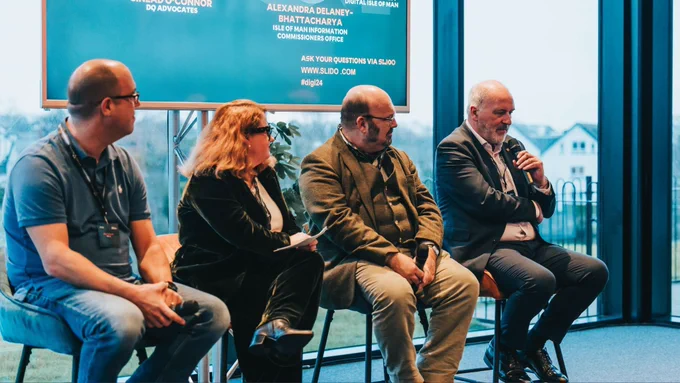 A photo of three men and a woman who are panellists for a discussion, with one man talking into a microphone