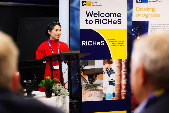 The speaker is looking out over the lecture theatre with an expression of pride. She stands beside a banner that reads 'Welcome to RICHeS'.