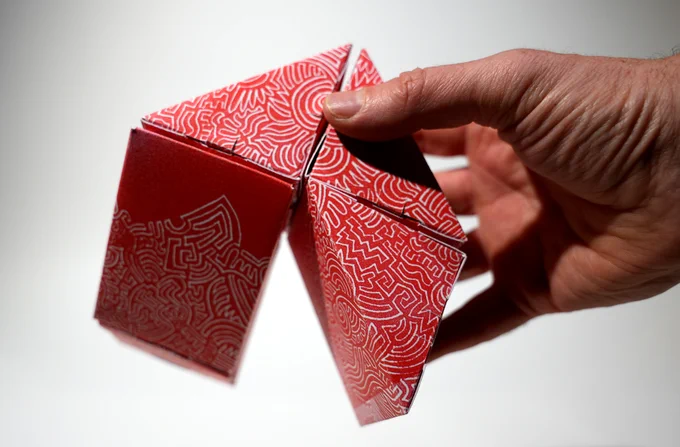 A hold holding an assembled Delightful Little thing. It consists of five triangular prisms made out of cardboard, interconnected to each other. It is red with a doodle linocut design.