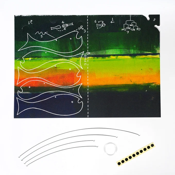 A Fish Mobile kit, which consists of: a colourful printed sheet of card with instructions and cutting guides, four wires, thread, and a trip of small round stickers. 
