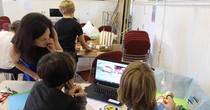 Two children, with backs to the camera, are working together on a stop-motion animation using a laptop. An adult is leaning over the table, looking at the laptop with them.
