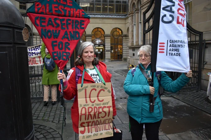 Two demonstrators, one with a Campaign Against Arms Trade flag and another with a placard reading "ICC Arrest Starmer".