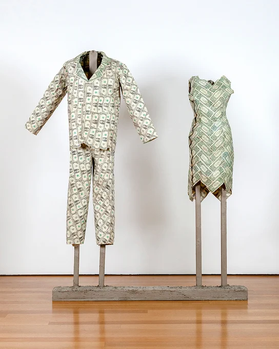 A suit and a dress made of hundreds of dollar bills displayed side by side on concrete armatures. On the men's suit jacket, the dollar bills flowing vertically down the body and pants. On the dress, the dollar bills criss-crossing down the garment like angled bricks.