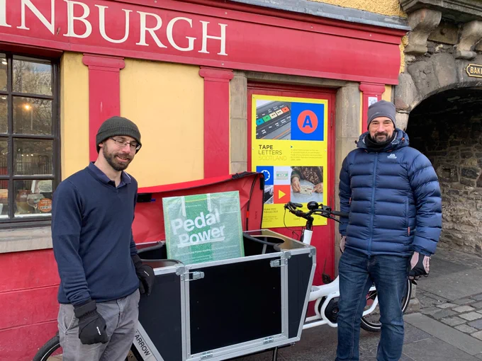 Two people standing by a cargo bike in front of the Museum of Edinburgh. The cargo bike is holding a display panel that reads "Pedal Power".