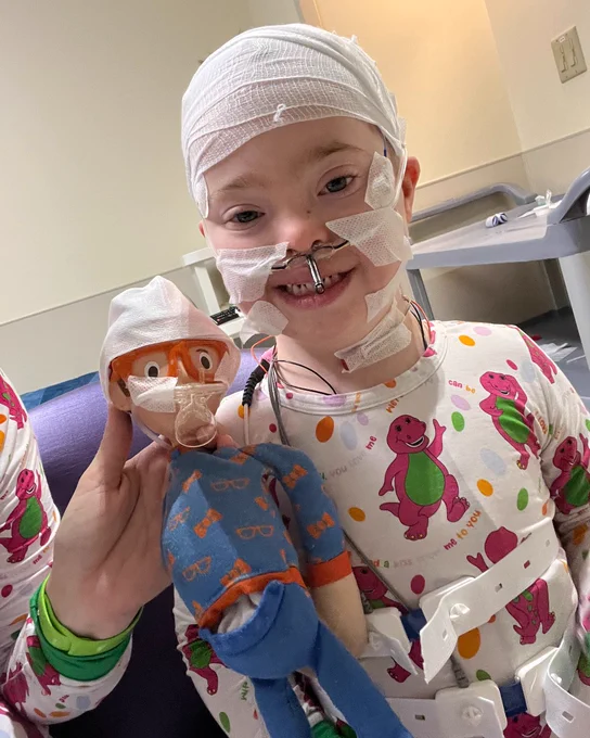 A child in hospital attire smiling and holding a stuffed toy version of Woody from Toy Story, both the child and the toy are wearing similar bandages around their heads.