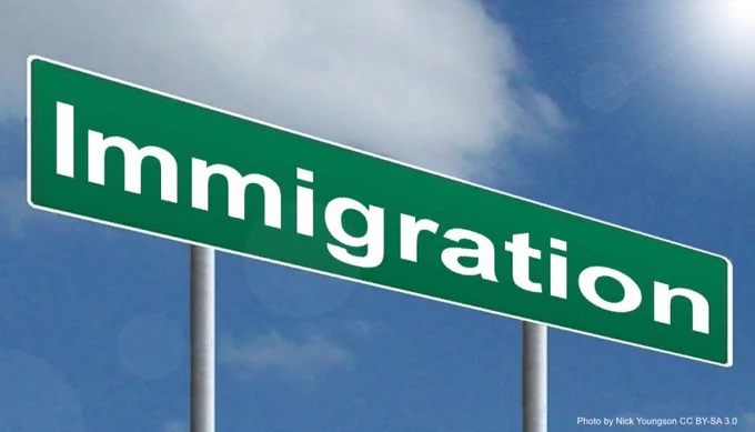 Green highway sign with the word "Immigration", against a blue sky.