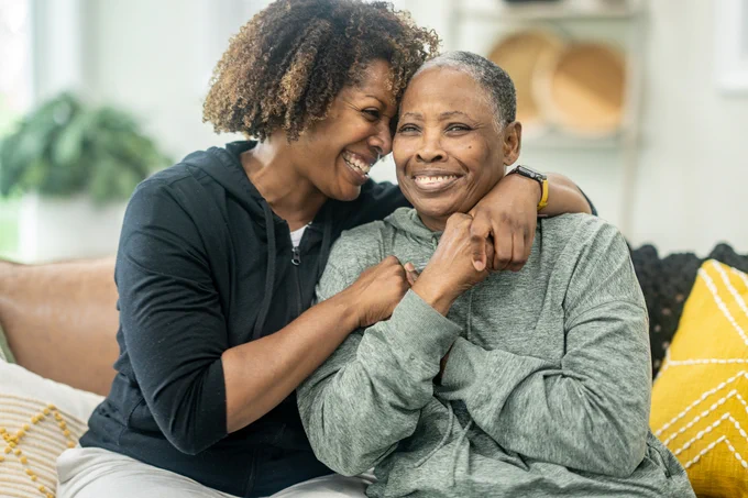 An African American woman wearing a black sweatshirt is hugging an elderly woman that is dressed in a light green sweater.