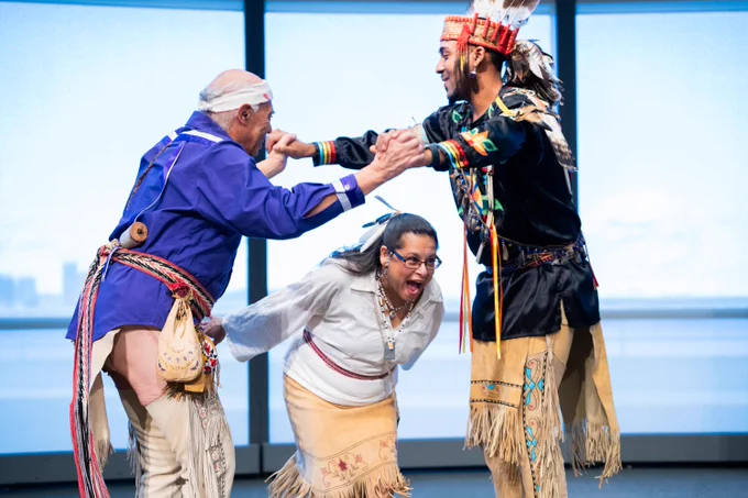 Three individuals in traditional Native American attire are performing a cultural dance at the JFK Library with large windows in the background.