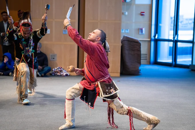 A member of the Wampanoag Nation in traditional attire is dancing energetically at the JFK Library, holding a feather aloft, with another member, also in traditional attire, in the background.