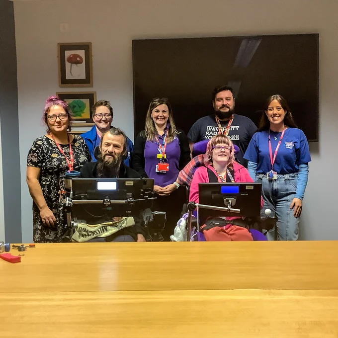 Image shows Henshaws College staff standing behind Jamie and Georgia, with everyone smiling at the camera. Jamie and Georgia are both in their chairs, with their chair screens in front of them. A classroom setting can be seen behind everyone.