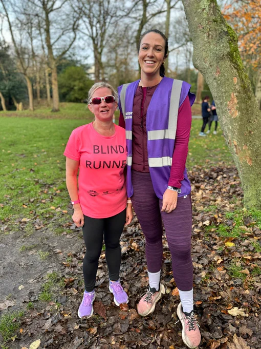 The image shows two women standing outdoors in a park-like setting with trees and grass in the background. The woman on the left is wearing a bright pink T-shirt with the words "BLIND RUNNER" printed on it, along with black leggings and purple running shoes. She is also wearing sunglasses. The woman on the right is taller and is wearing a purple high-visibility vest over a maroon long-sleeve top, maroon leggings, and pink running shoes. They are both smiling and standing on a path covered with fallen leaves.