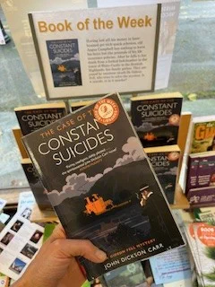 Image is a book held by a hand. The cover depicts a man standing by the side of a lake smoking a cigar and looking at a large house on the other side of the lake