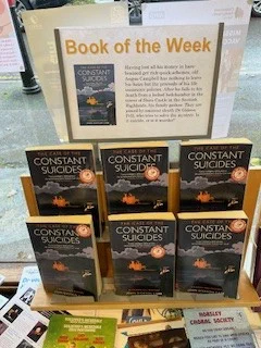  Image is a photograph of Horsley Library's Book of the Week display, a plastic stand featuring 6 copies of John Dickinson Carr's "The Case of the Constant Suicides". Above the display is a laminated advertisement in landscape format. Said advertisement is titled: 'Book of the Week' and has a picture of the Book's cover on the left and the text of the blurb on the right, as in the body of the post.
