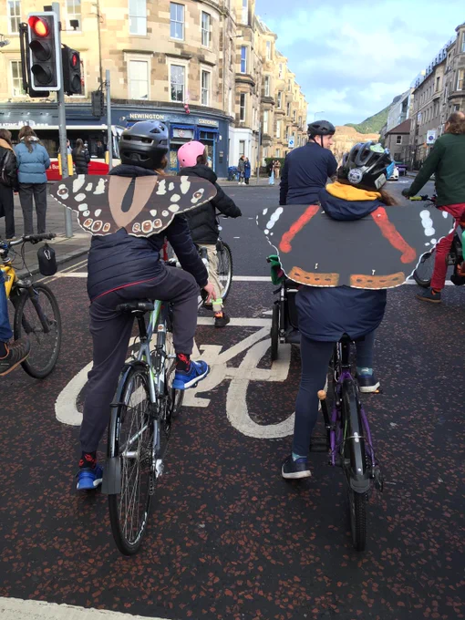 Two people wearing cardboard butterfly wings sitting on their bikes, stopped at a traffic light in Newington. Around them are several more cyclists.
