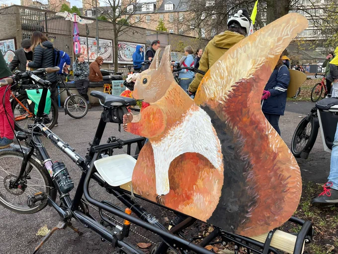 A cargo bike with a cardboard red squirrel in the cargo bay. Around the bike are many other bikes and cyclists.