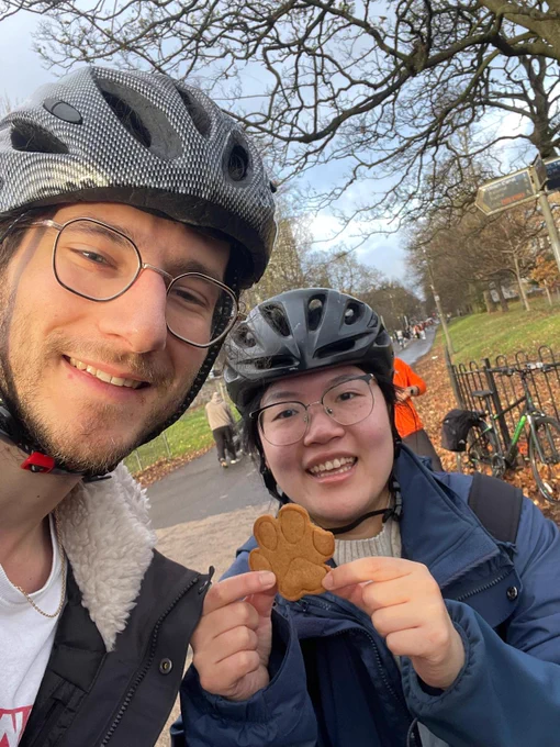 Two people in bike helmets wearing jackets. They are standing in a park. One is holding a paw-shaped biscuit.