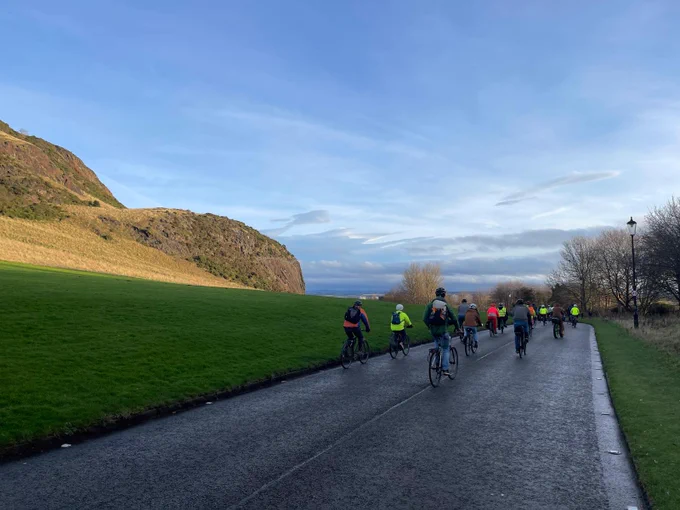 Several cyclists riding on a road in Holyrood Park. Behind them is a big hill.