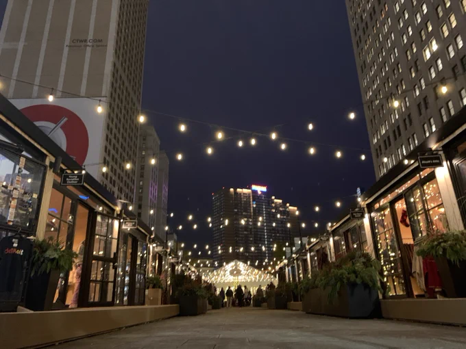 An evening view of a city walkway lined with shops and cafes, adorned with string lights overhead, set against a backdrop of tall buildings.
